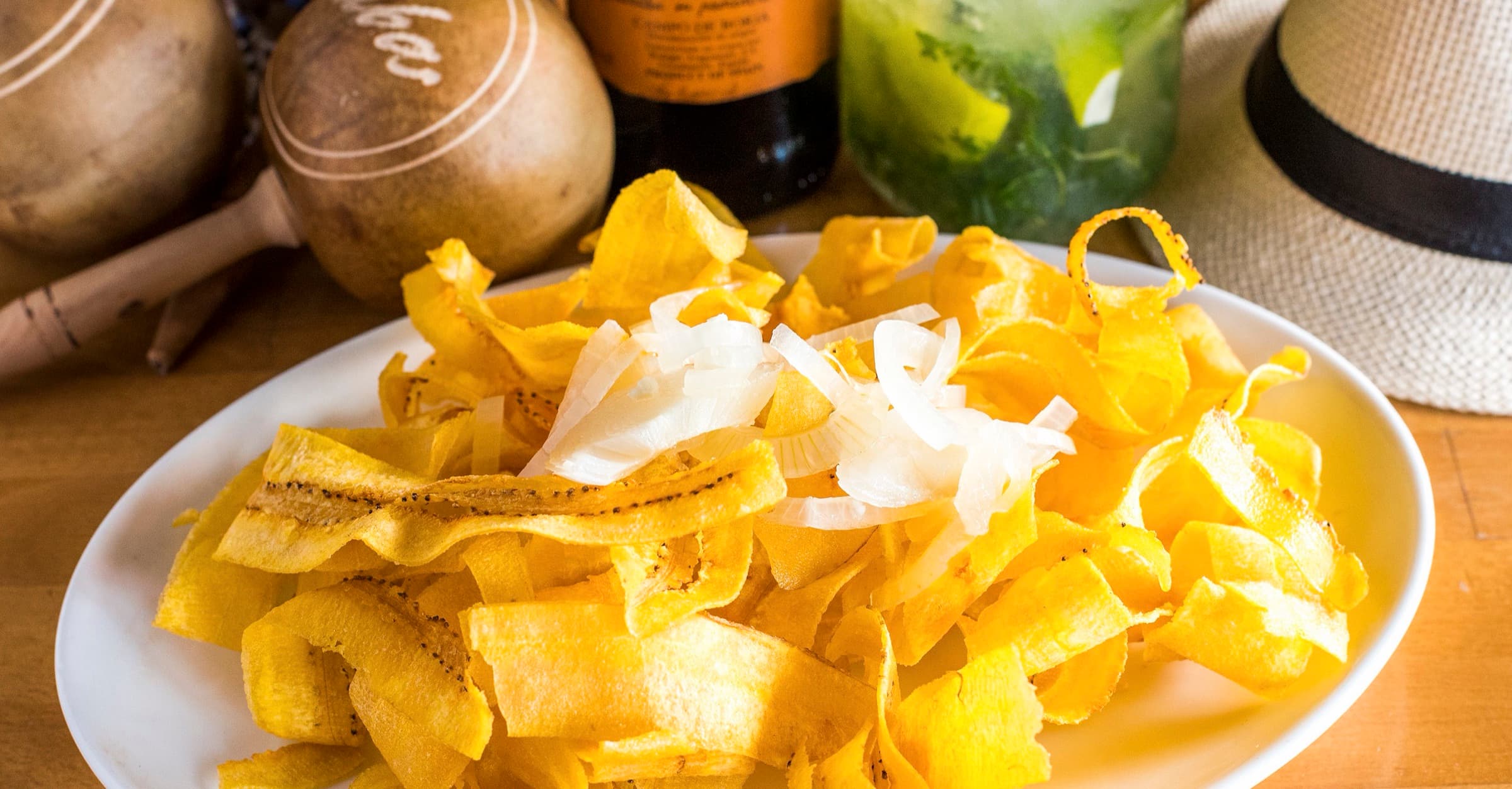 A platter of chicharritas topped with raw onion, beside maracas, a mojito and a straw hat